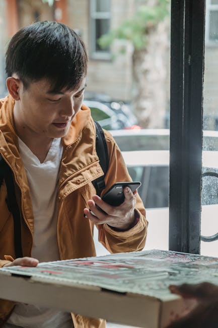 Asian man delivering pizza outdoors, holding a smartphone, urban setting.
